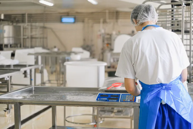 Técnica de touca e avental a pesar numa balança digital, numa cozinha de produção em inox